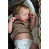 A newborn Reborn baby doll with rooted hair sleeping in a woven basket surrounded by soft fabric.