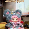 A fluffy blue bear plush charm with pink accents and a dark blue cape is displayed on a wooden surface.