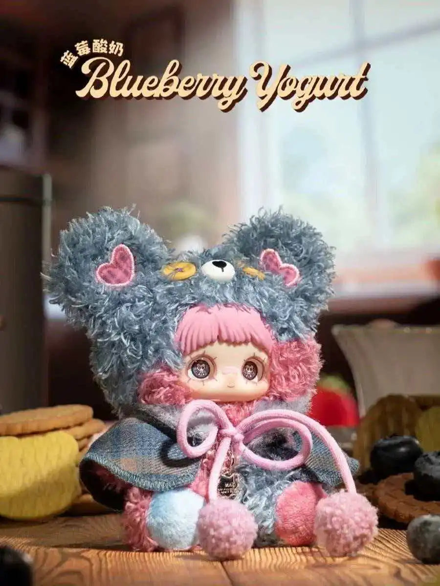 A fluffy blue bear plush charm with pink accents and a dark blue cape is displayed on a wooden surface.