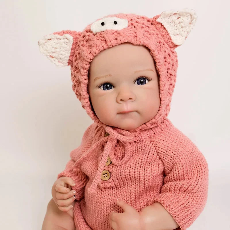 A realistic silicone reborn baby doll wearing a pink knitted animal hood and sweater.