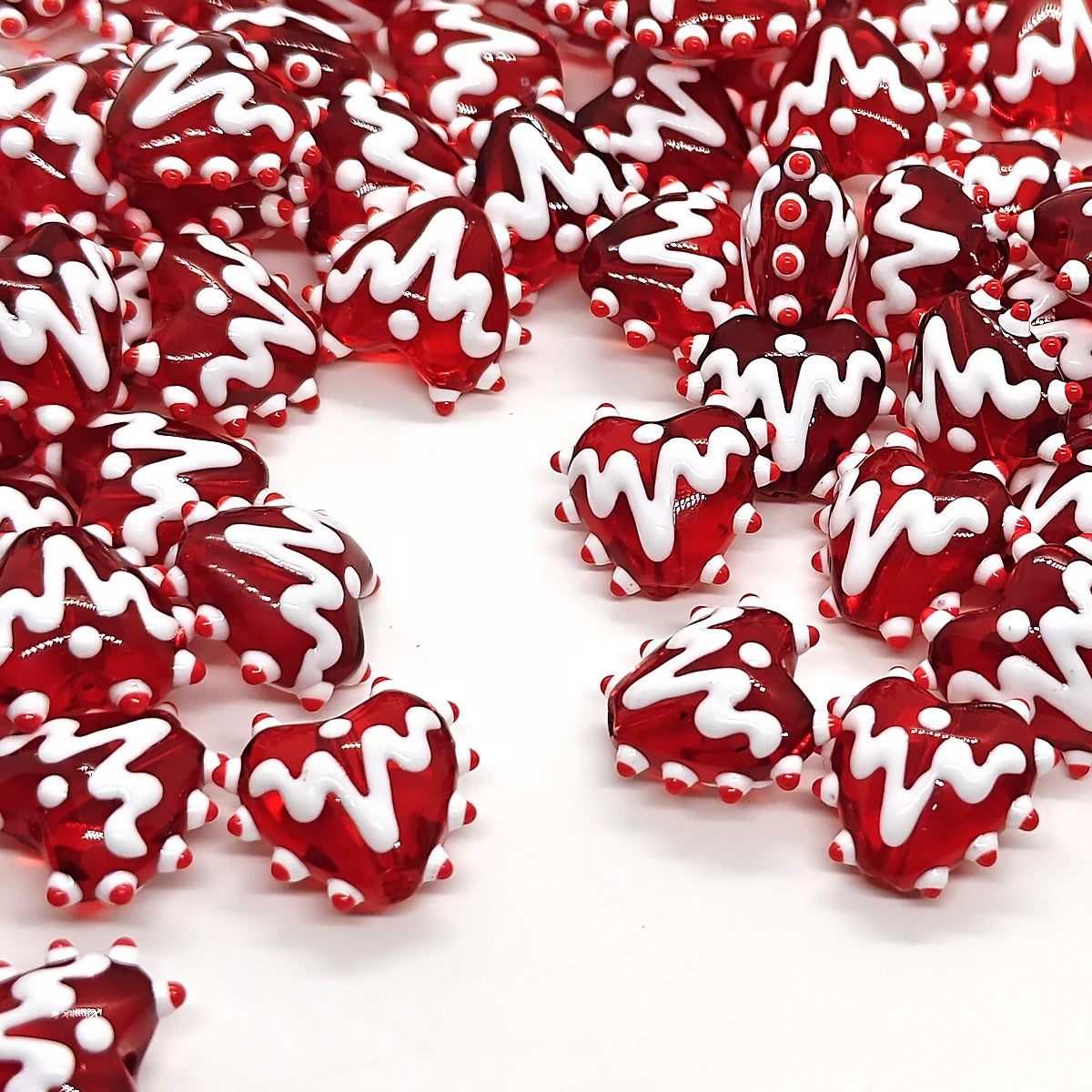 A close-up of red heart lampwork glass beads with white swirling designs scattered on a white background.