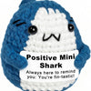 A blue and white crocheted shark plush toy with a happy expression and a tag that reads Positive Mini Shark You're fin-tastic
