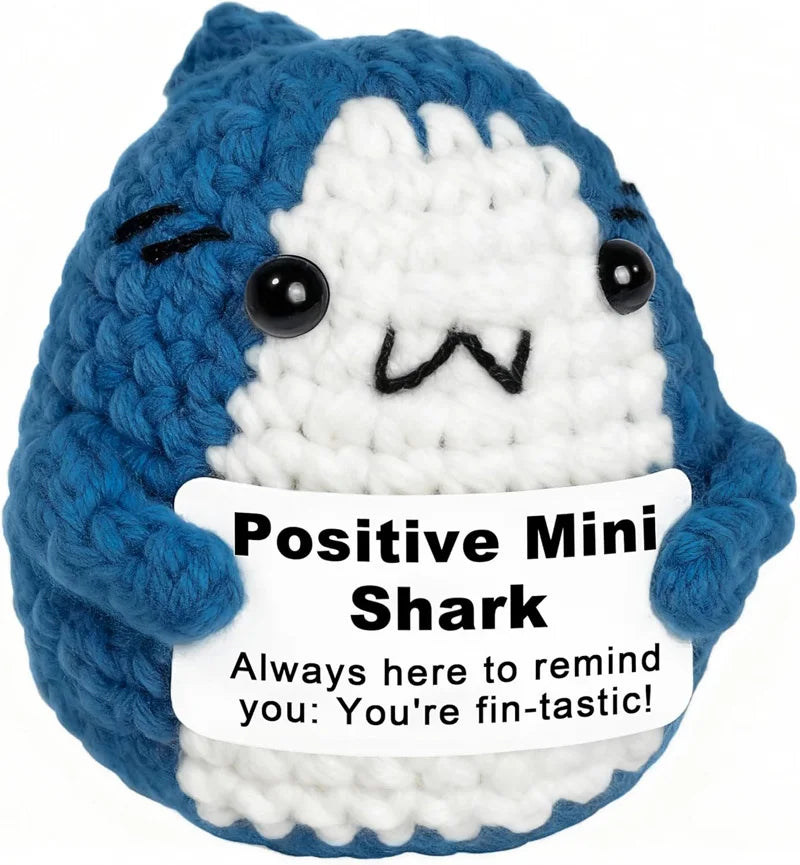 A blue and white crocheted shark plush toy with a happy expression and a tag that reads Positive Mini Shark You're fin-tastic