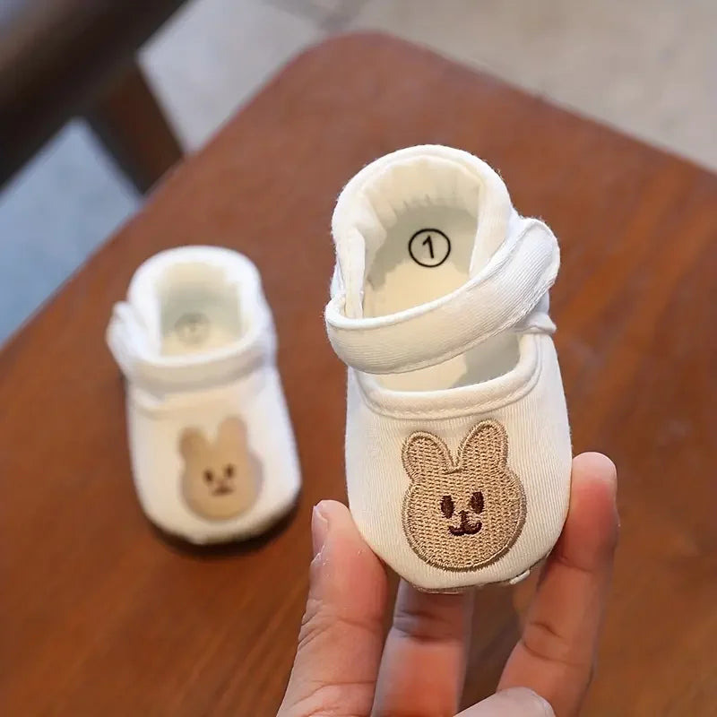 A pair of white soft sole first walker shoes shaped like bears, shown being held by a hand.