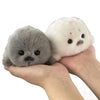 A small grey seal plush toy is held in a person's hand against a white background.