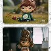 A small mischievous figure with brown hair and green skin sits on grass, displayed next to a teddy bear figure.