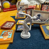 A white figurine of a leaping figure is displayed among various framed miniature paintings on a blue fabric surface.