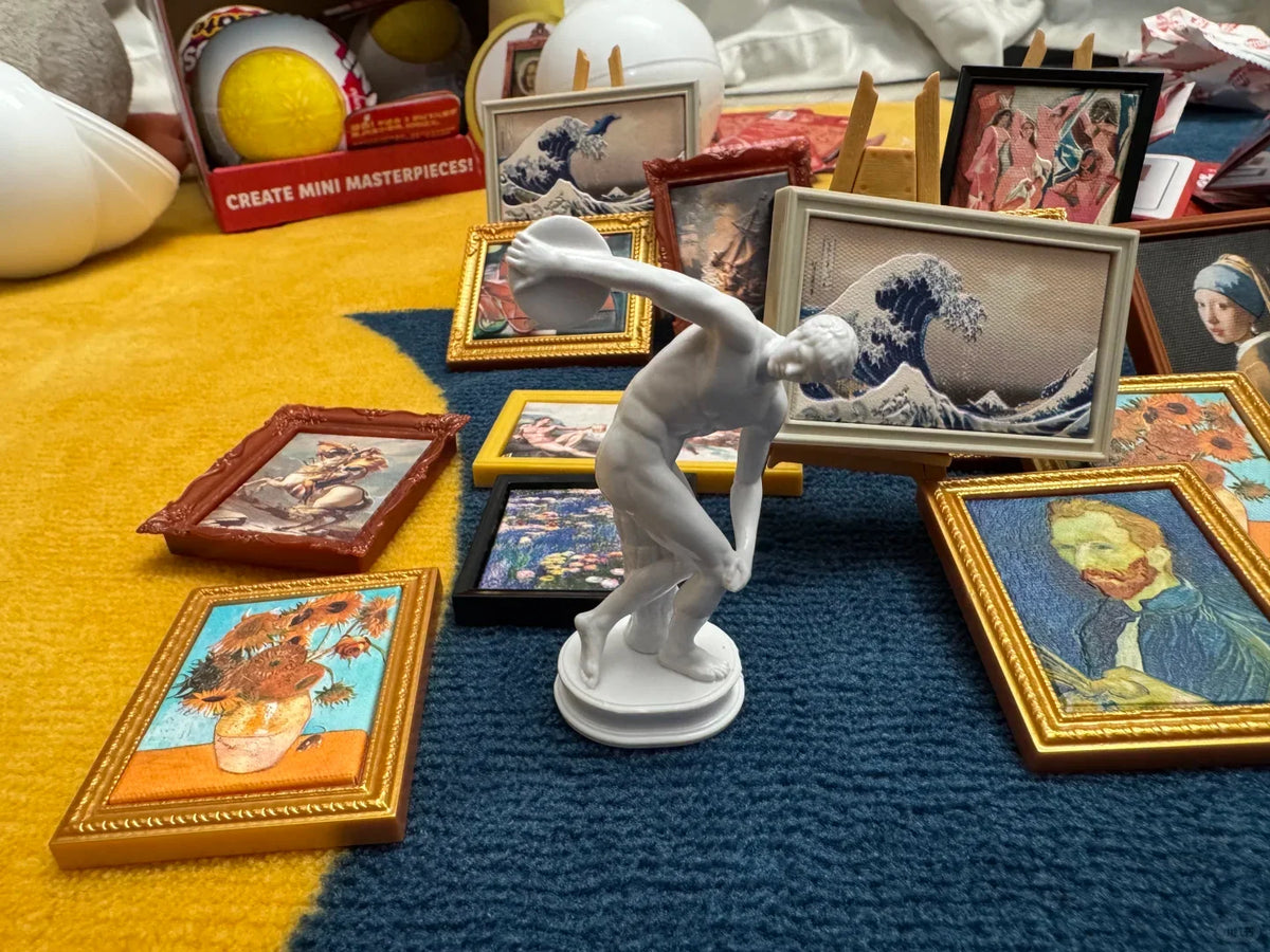 A white figurine of a leaping figure is displayed among various framed miniature paintings on a blue fabric surface.