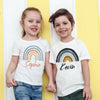 Two children, a girl and a boy, stand side by side wearing white t-shirts with rainbow designs and names.