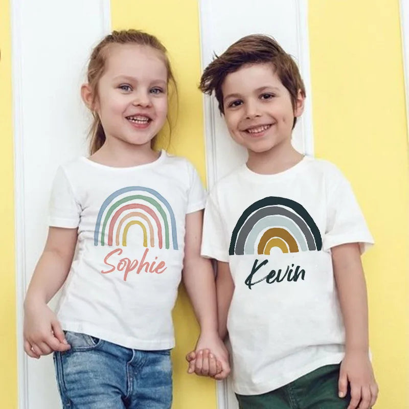 Two children, a girl and a boy, stand side by side wearing white t-shirts with rainbow designs and names.