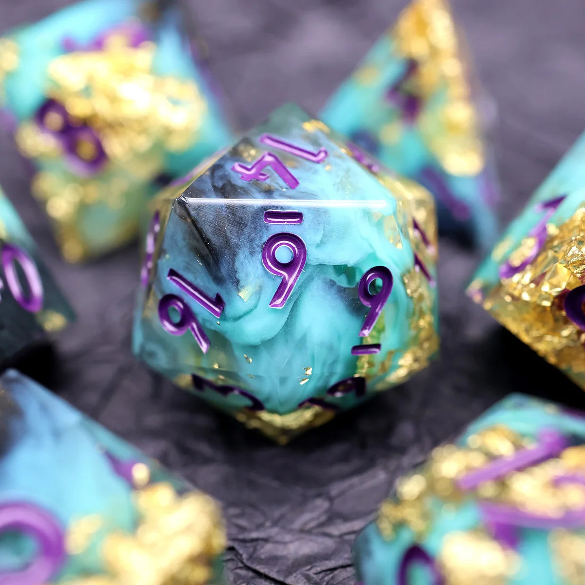 A close-up of several teal resin polyhedral dice with gold accents and purple markings resting on dark fabric.