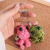 A hand holds a pink and green turtle plush keyring charm with silver jump rings on a brown cork board.