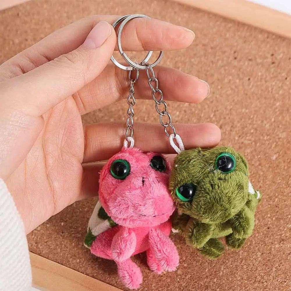 A hand holds a pink and green turtle plush keyring charm with silver jump rings on a brown cork board.