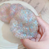 A hand holds a clear, iridescent squishy stress relief ball with blue and pink hues on a white surface.