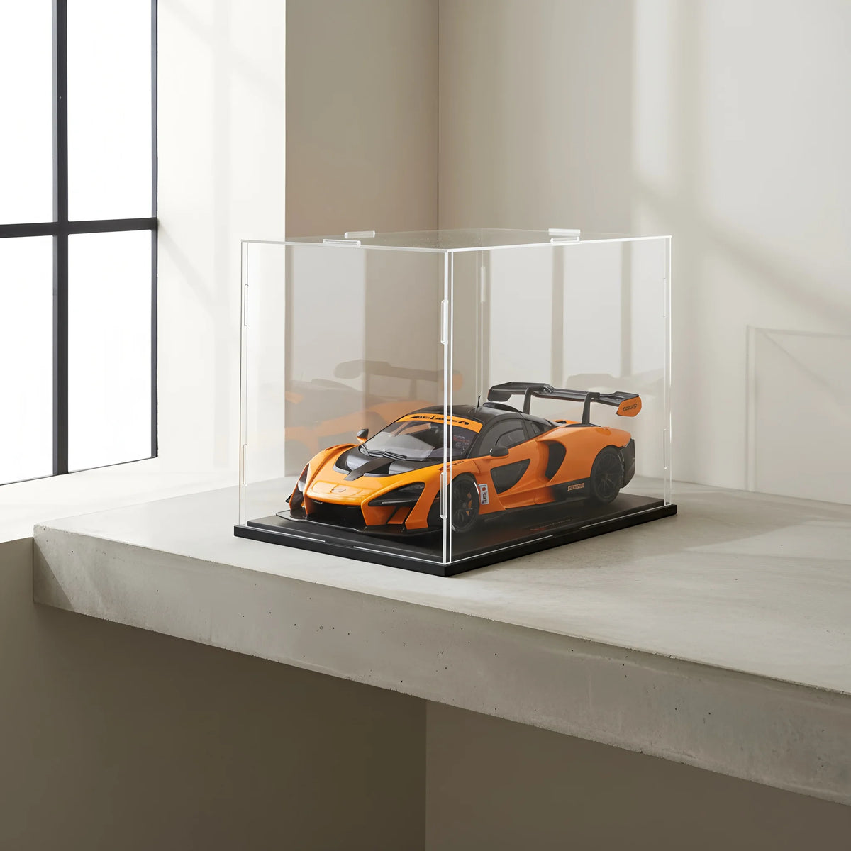 Une voiture de sport orange est exposée dans une boîte en acrylique transparente sur une surface claire.