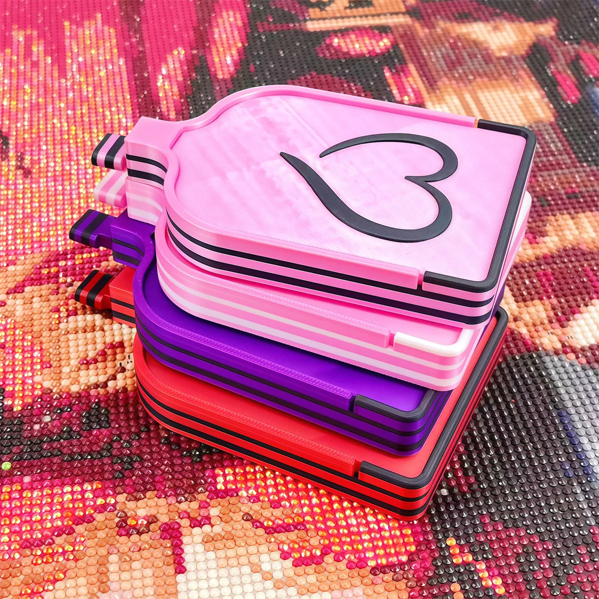 A stack of pink, purple, and red diamond painting storage trays and organizer boxes on a textured background.