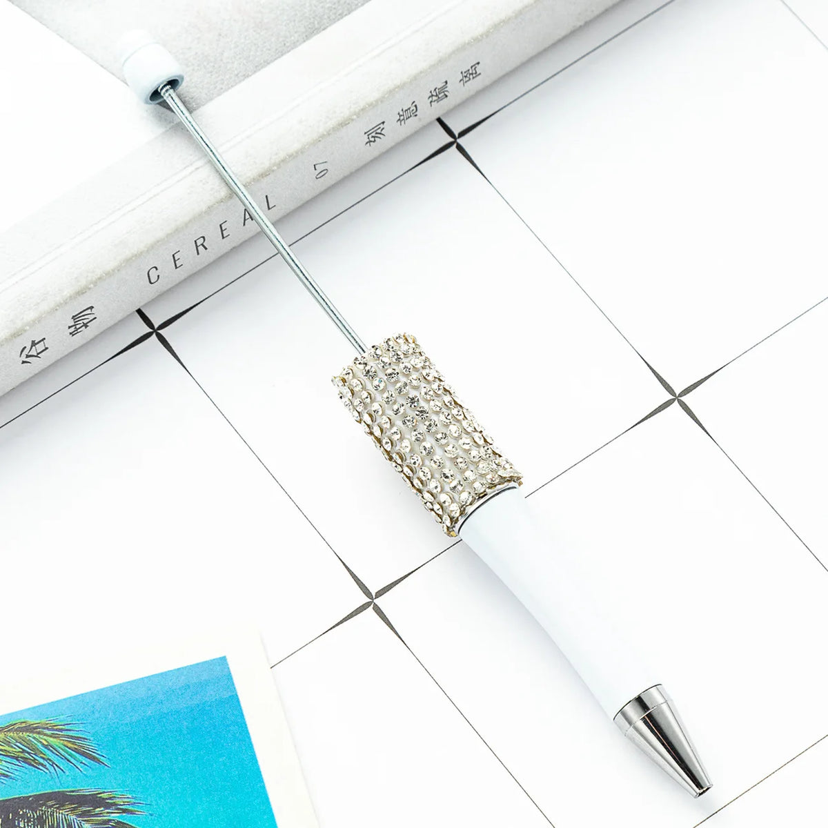 A white ballpoint pen with a silver beaded tip rests on a white surface next to a notebook.