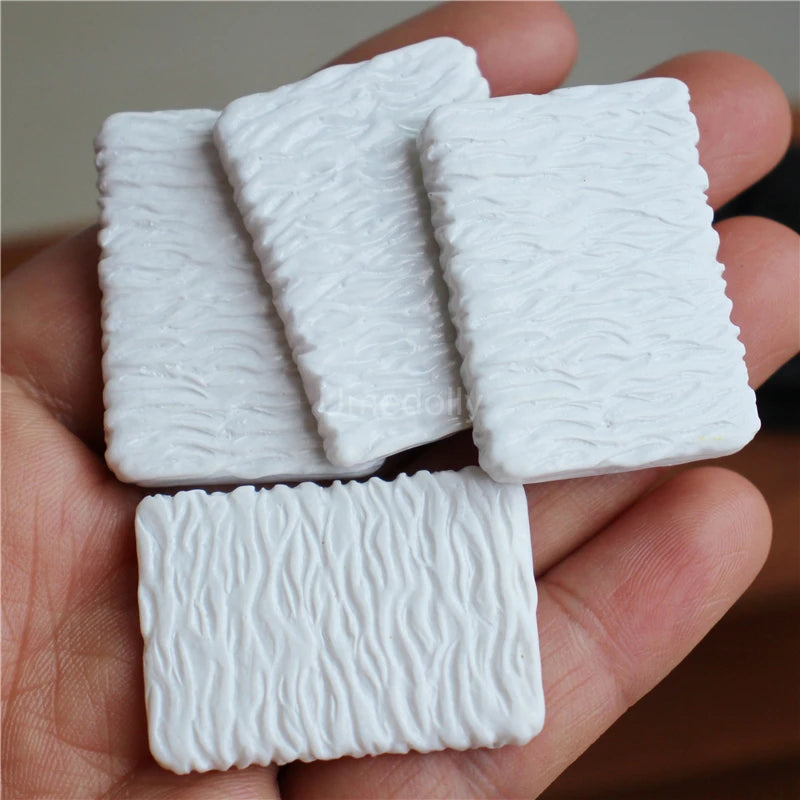 A hand holds four rectangular white miniature food items, likely a dollhouse set for beef steak and pasta.