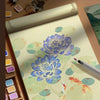A watercolor paint set featuring blue flowers and a goldfish on textured paper, displayed with brushes and paint pots.