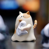 A small, light-colored figurine of a cat gently holding a white, blob-like object.
