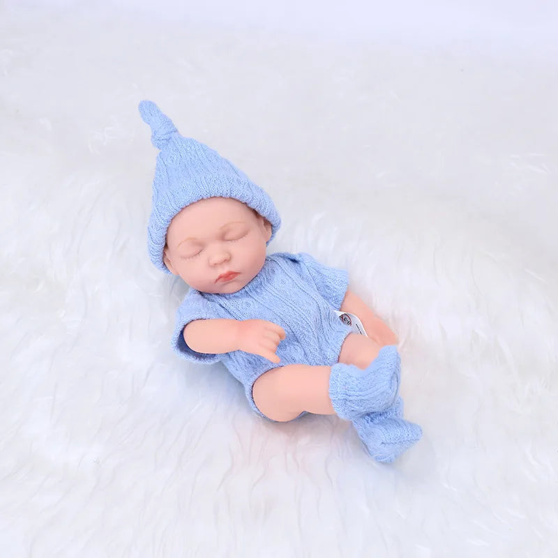 A realistic soft vinyl baby doll wearing a blue knitted hat and matching outfit, lying on a white surface.