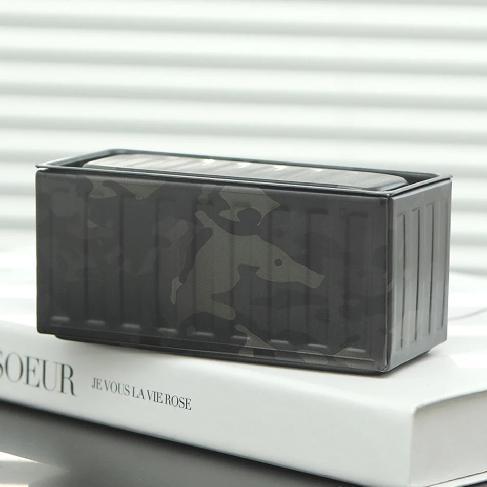 A dark camouflage miniature industrial iron storage box with an openable lid resting on a white book.