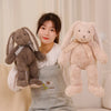 A young woman holds a light beige bunny plush toy with long ears and a bow in a soft indoor setting.