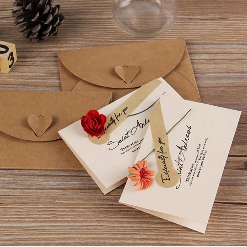 A set of kraft paper envelopes with heart clasps and a red rose decoration are displayed on a wooden surface.
