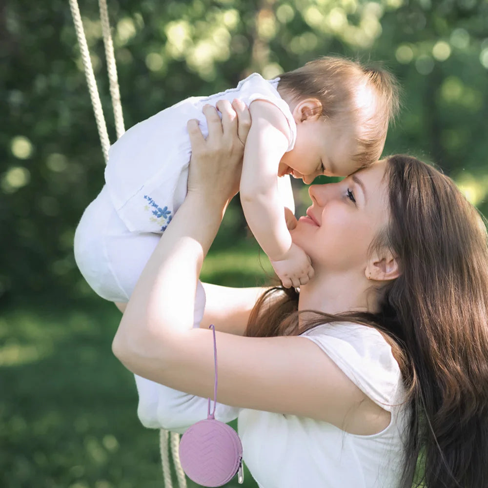 A young woman holds a baby in a sling while using a silicone pacifier holder and soother case outdoors.