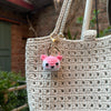 A handcrafted crochet animal keychain shaped like a pink and white rabbit is attached to a beige woven tote bag.