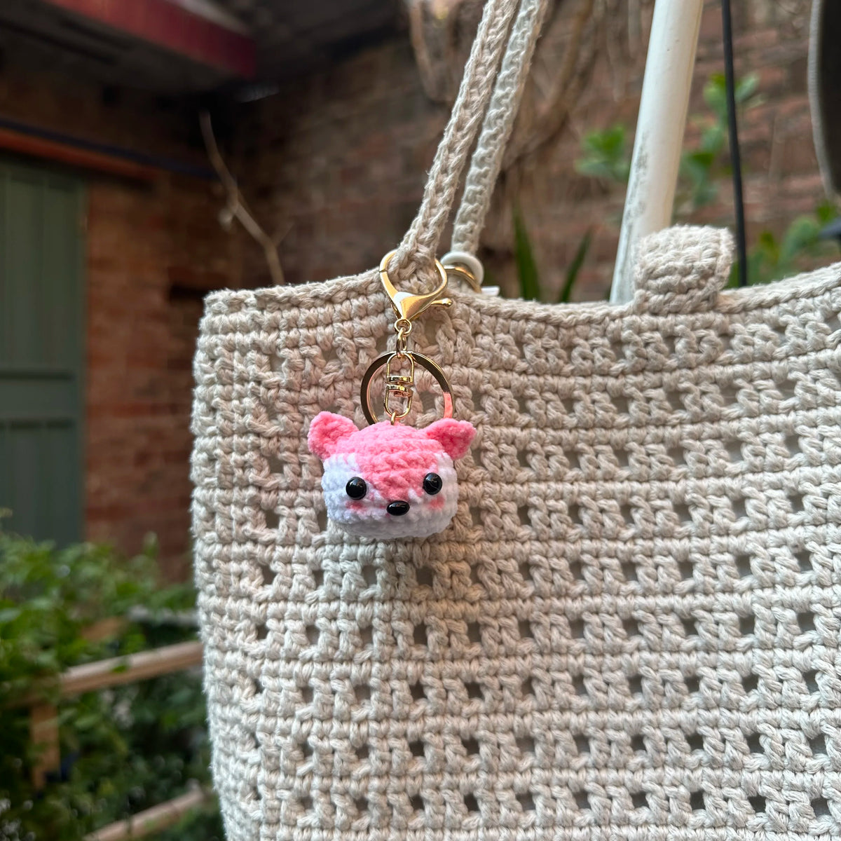 A handcrafted crochet animal keychain shaped like a pink and white rabbit is attached to a beige woven tote bag.