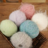 A collection of fluffy pink, white, and light blue long-haired alpaca blend yarn balls resting on a woven tray.