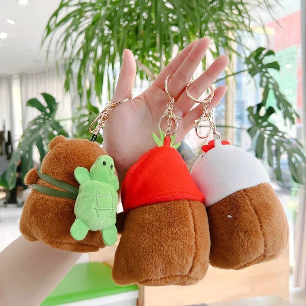 A hand holds three adorable capybara plush keychain charms in shades of brown, green, and white.