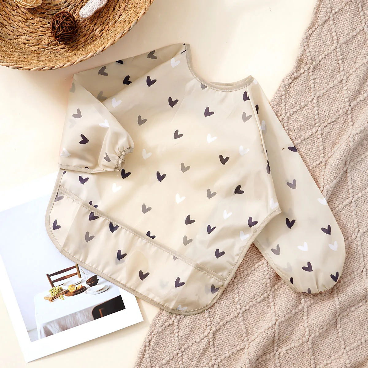 A beige waterproof long sleeve feeding bib with a subtle pattern of gray and black hearts is displayed on a textured fabric.