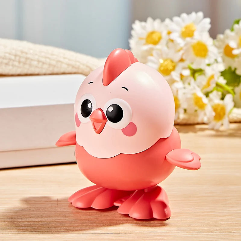 A pink wind-up chick toy with a pull chain sitting on a wooden surface next to white boxes and white flowers.