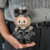 A small, fluffy doll wearing a black and white flannel outfit with a bow is held in a person's hand.