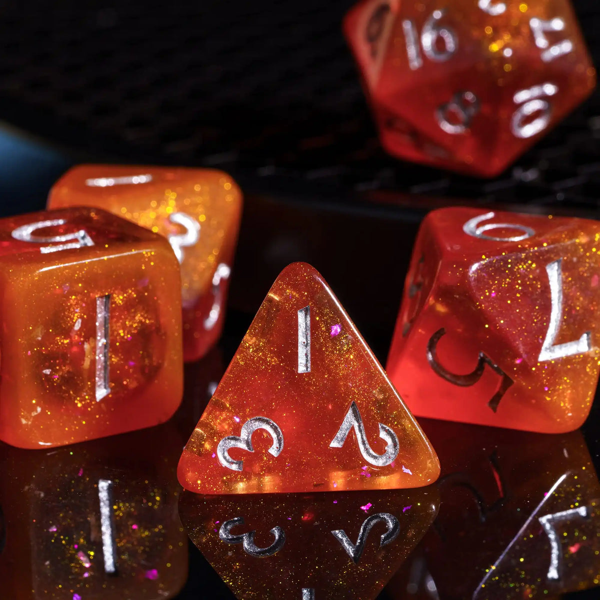 A close-up of several orange and red glitter resin polyhedral dice resting on a dark surface.