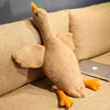 A fluffy, tan plush goose toy with orange feet is resting on a beige sofa next to an Apple laptop.