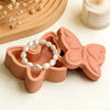 A terracotta silicone resin mold shaped like a butterfly holds a strand of white pearl beads.