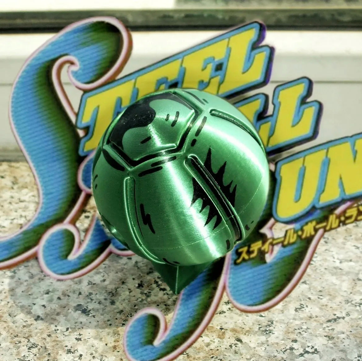 A green steel ball fidget toy with a stylized yellow and blue logo in the background.
