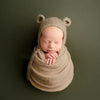 A newborn baby is swaddled in a beige knit bear hat and wrap set against a dark green background.