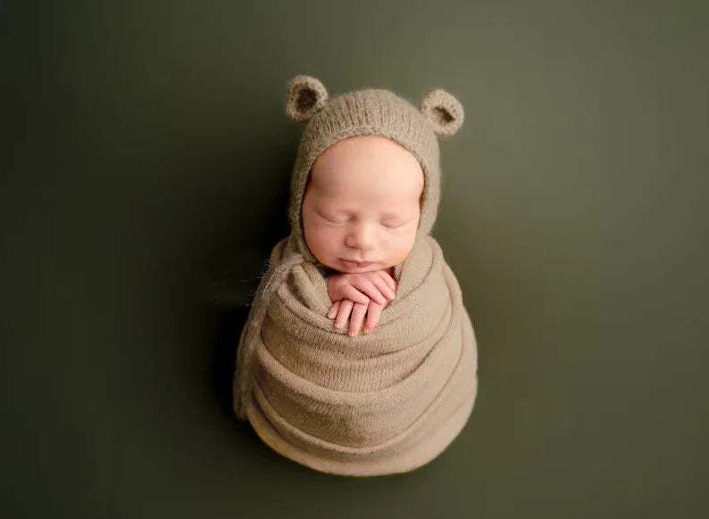 A newborn baby is swaddled in a beige knit bear hat and wrap set against a dark green background.