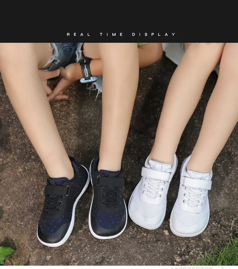 A close-up of a child's feet wearing dark blue and black barefoot training sneakers on a natural ground surface.