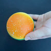 A hand is holding an orange squishy hamburger stress ball with white speckles against a dark background.