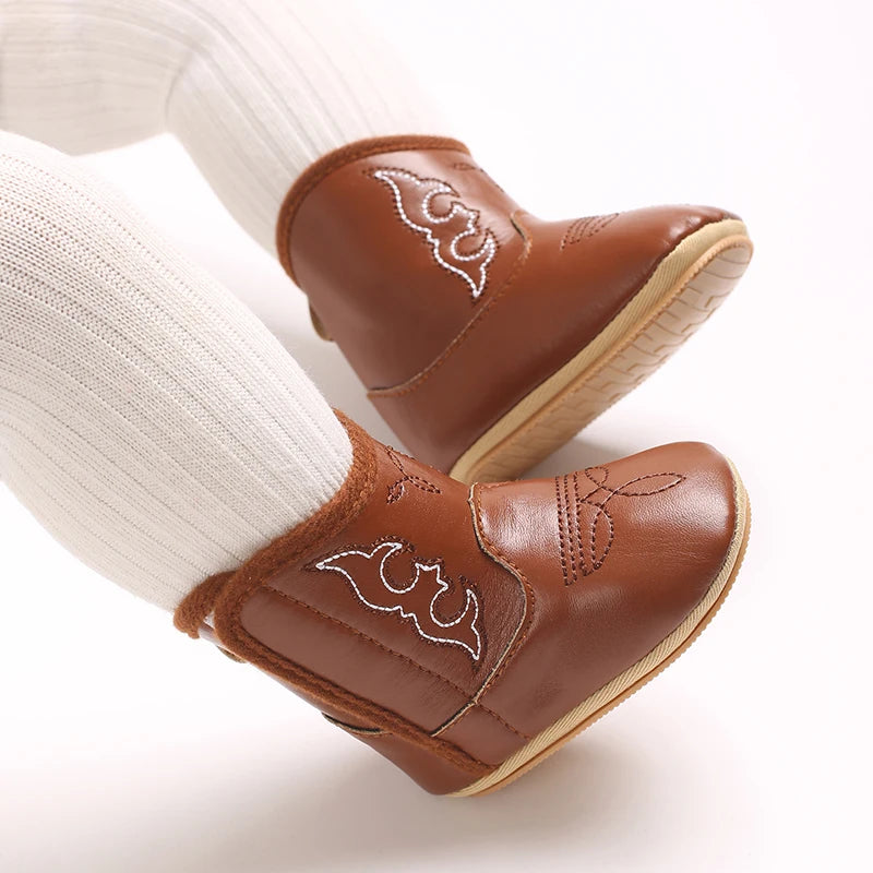 A pair of brown baby western booties with white stitching and a soft non-slip sole, shown on a white leg.