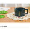 A dark ceramic coffee cup sits on a light wooden surface next to a white crocheted doily.