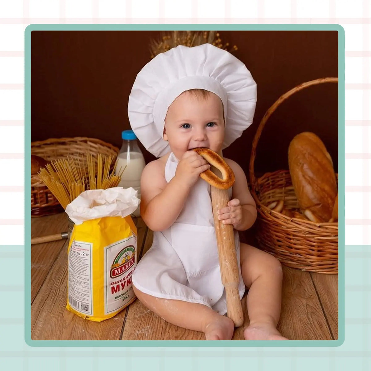 A baby wearing a white chef hat and apron is sitting next to a loaf of bread and a bag of flour.
