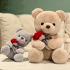 A large tan teddy bear plush toy holding a red rose, next to a smaller gray teddy bear.