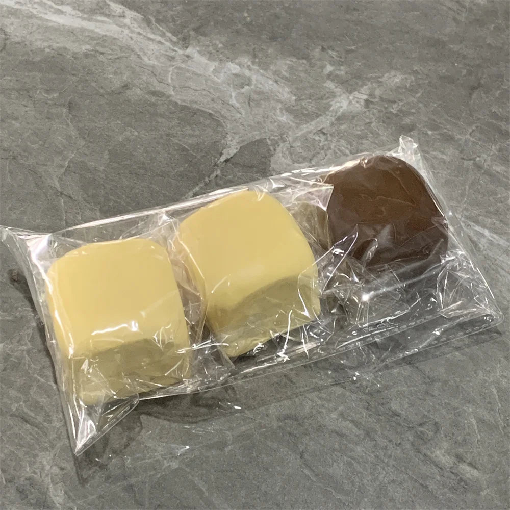 Un sac plastique transparent contient deux jouets de pains décoratifs jaunes et un jouet brun sur une surface grise.