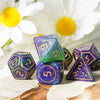 A set of seven purple and green polyhedral resin dice displayed on a wooden surface with white flowers in the background.